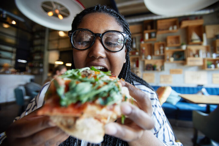 Hungry young woman in eyeglasses going to bite piece of appetizing pizza by lunch in cafe