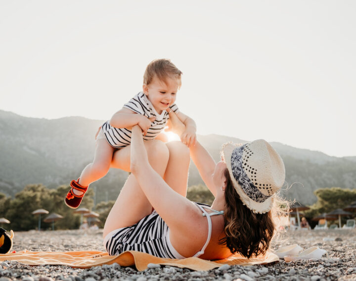Young mother having fun with her little daughter sitting on the beach in the mountains during summer sunset. Happy family concept. Happy childhood.