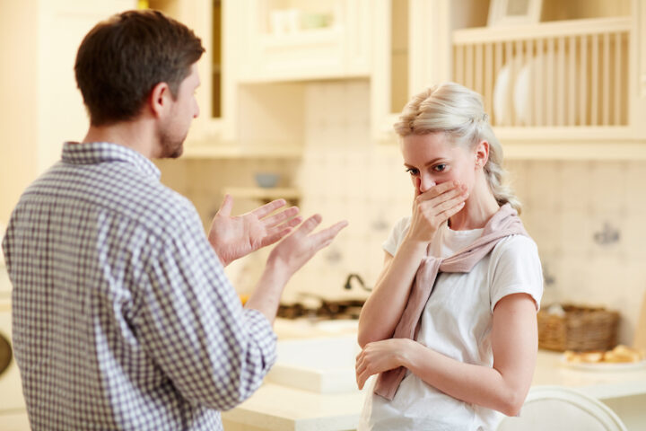 Upset young woman covering her mouth and expressing displeasure while irritated husband talking to her in rude manner