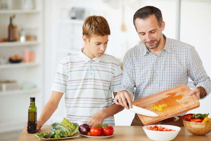Young father cooking vegetable salad and teaching his son to cook in domestic kitchen