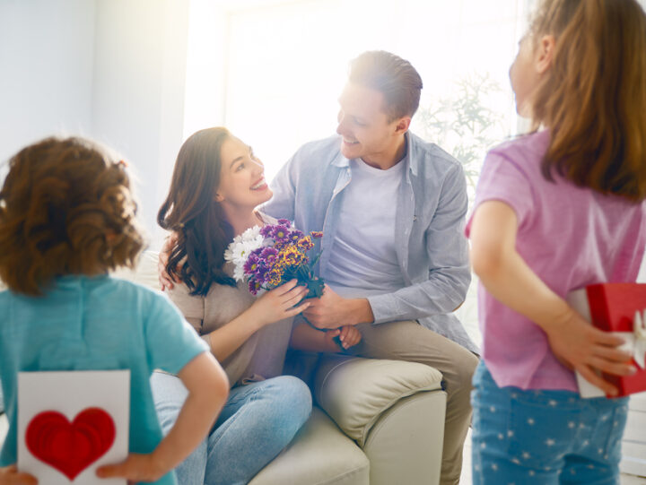 smiling dad giving flowers to his wife, while two kids wait hiding a gift and a card behind their back