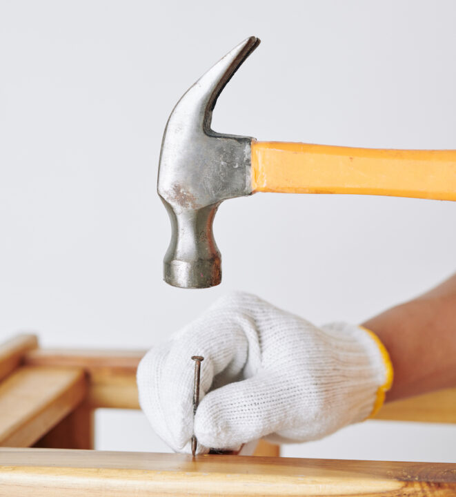 Close-up image of carpenter in textile gloves hammering nail when making stool in his workshop