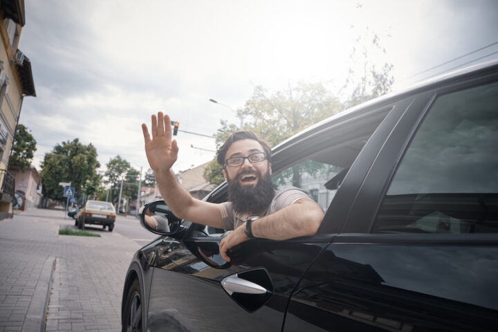 Happy man in the passenger seat of a car, waving through the opened window, saying hello