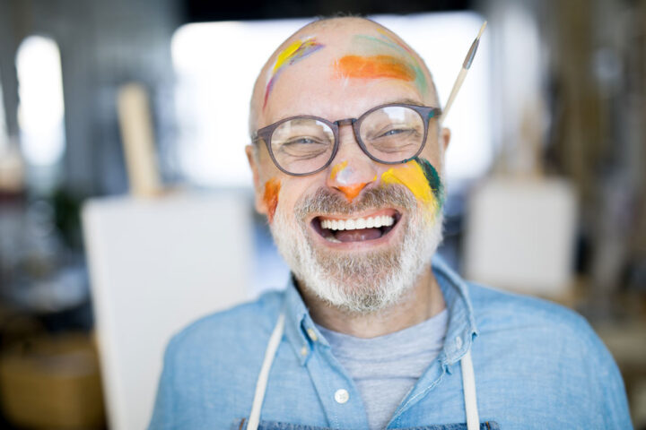 Bald mature artist with grey beard and paints all over his face looking at camera