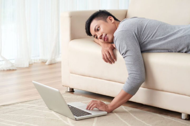 Man lying on the couch and looking at his laptop with a lazy expression
