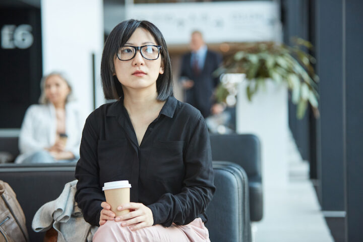 Sad young Asian woman wearing black shirt sitting alone in airport departure lounge holding cup of coffee looking away