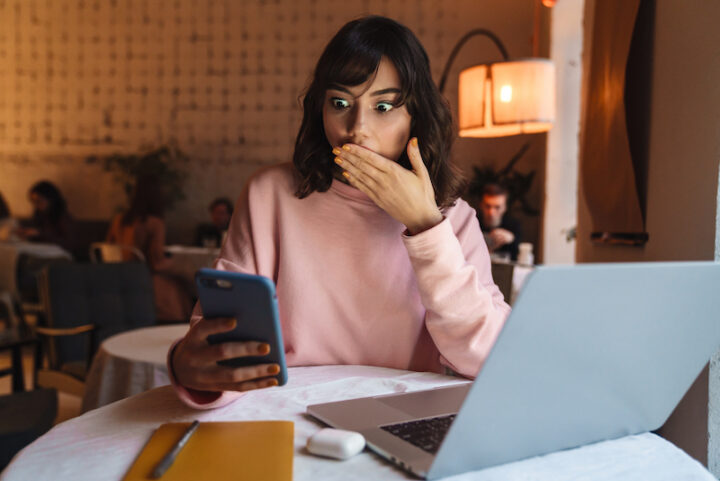 Photo of a beautiful shocked woman indoors in cafe using laptop computer and mobile phone.
