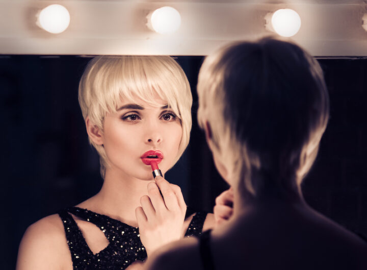 Beautiful Blonde Woman Looking Into The Mirror At Herself And Applying Red Lipstick.