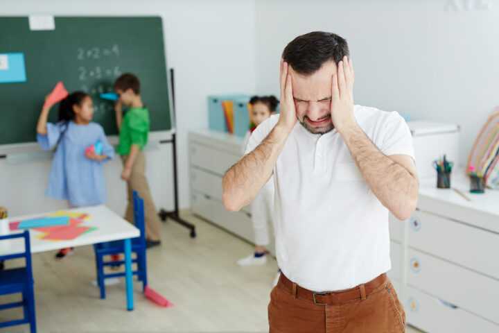 Annoyed teacher touching his head during break between lessons while group of kids having fun on background