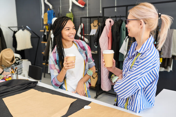 Two young female tailors smiling and talking to each other while standing and drinking coffee in workshop