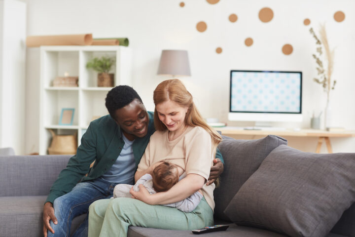 Portrait of happy interracial family sitting on couch at home together with mature mother breastfeeding baby, copy space