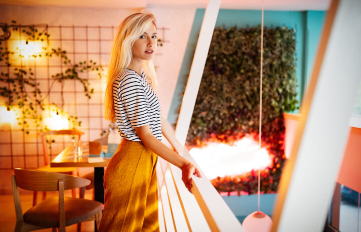 young blonde girl standing in a indoor balcony of a cafe, looking at the camera