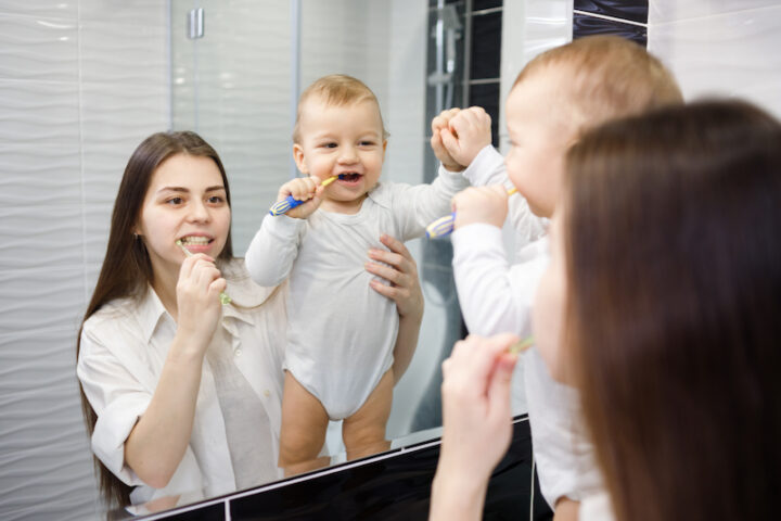 Young caucasian mother teaching baby boy how to brush teeth with toothbrush.