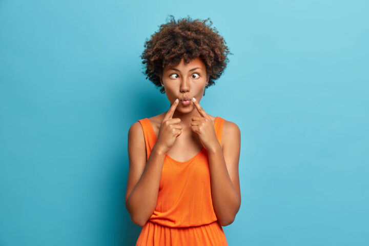 Funny dark skinned woman crosses eyes and makes fish lips funny grimace dressed in fashionable dress isolated on blue background.