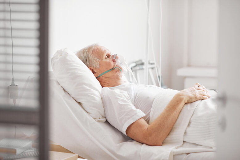 Side view of sick senior man lying in hospital bed, wearing oxygen supplement mask, eyes closed, copy space