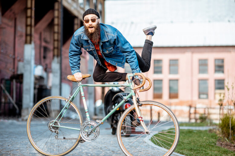 A bearded man is a crazy hipster playing with a retro bicycle outdoors against an industrial city background