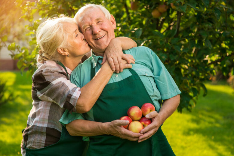 Elderly couple with apples, woman kissing man outdoors.