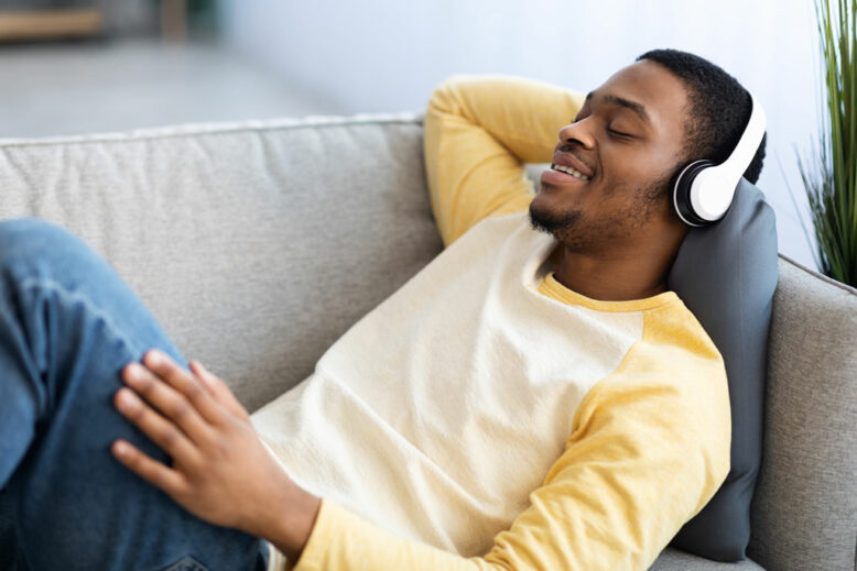 African american guy music lover laying on couch at home, listening to music and singing, using brand new wireless headset