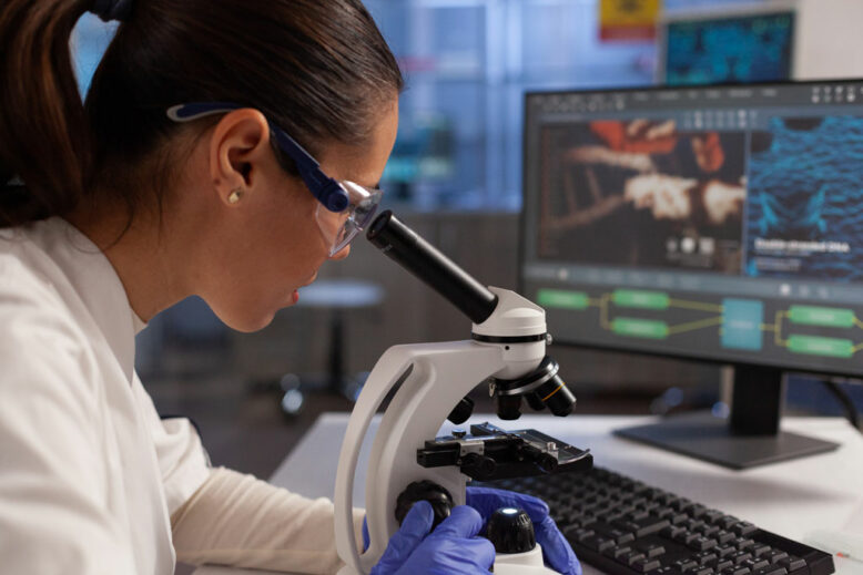 Biochemist woman working on microscope for cell illustration on computer screen in chemical laboratory.
