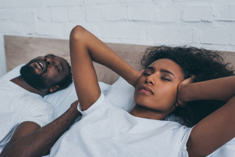 exhausted african american woman covering ears with hands while lying near snoring husband