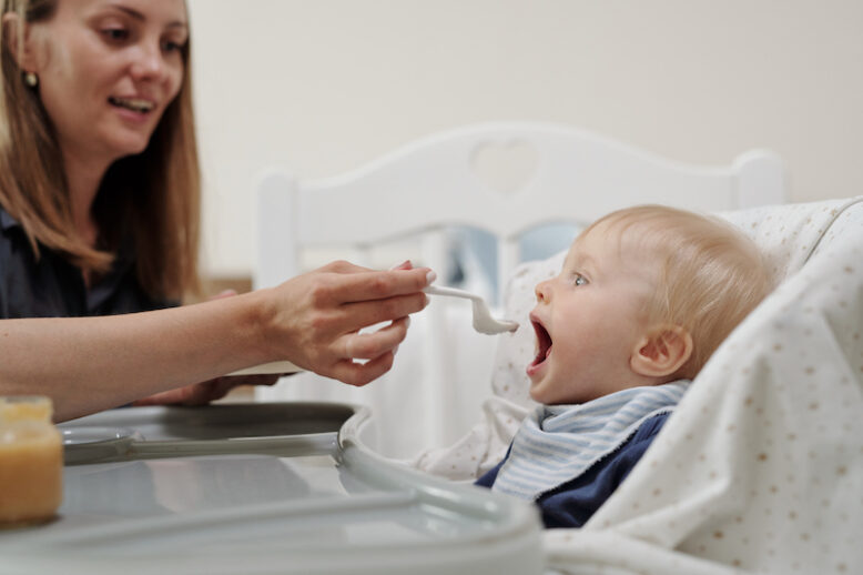 Positive young mother giving spoon of baby food to son sat in highchair while having lunch