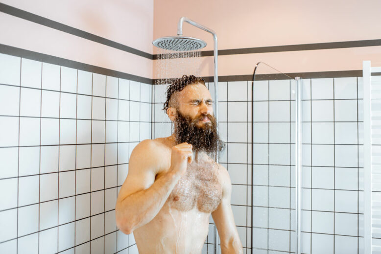 Bearded man feeling shocked while taking a shower with cold water in the bathroom at home