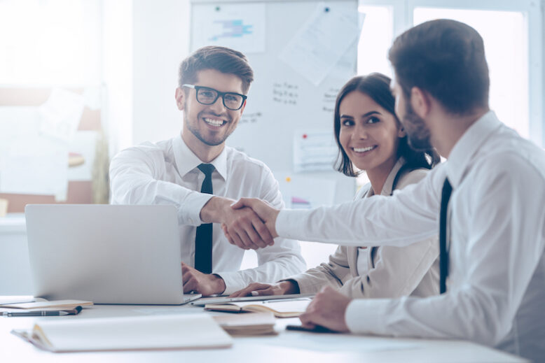 Two man shaking hands with smile while sitting at the office table with their coworker