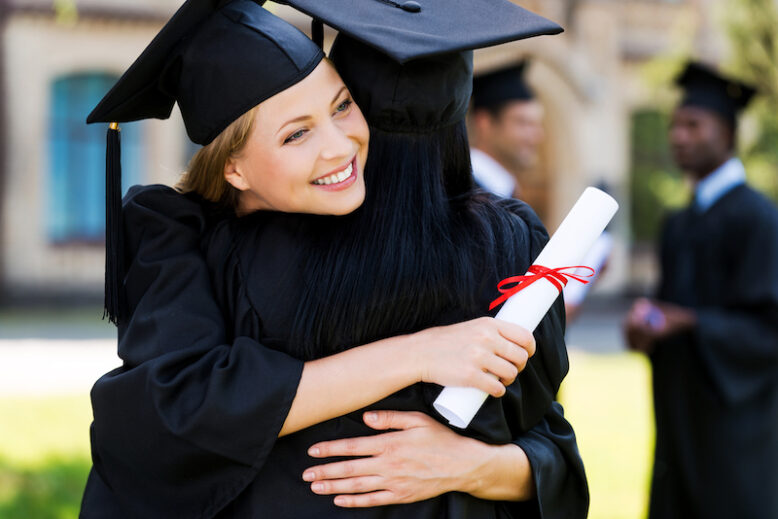 Two happy woman in graduation gowns hugging and smiling
