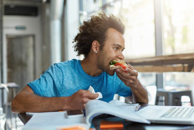 Black mixed race male student being busy with studying resting for minute eatting sandwich.