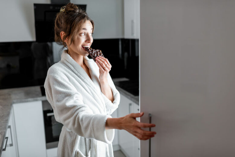 Young woman in bathrobe eating chocolate near the fridge, feeling hungry at night.