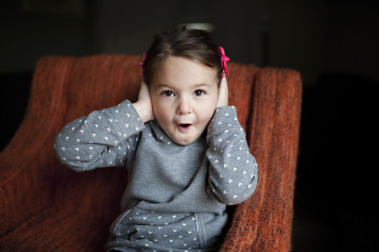 young girl sitting on a red arm chair covering her hears