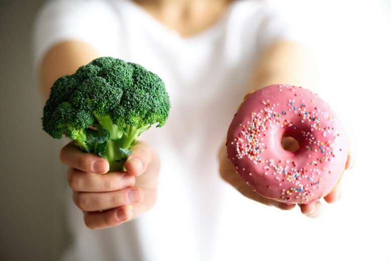 Young woman in white T-shirt choosing between broccoli or junk food, donut.