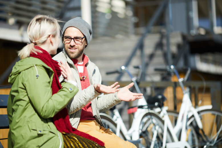 Young caucasian bearded man wearing glasses chatting with his girlfriend while sitting on a bench next to his parked bicycle on a sunny autumn day