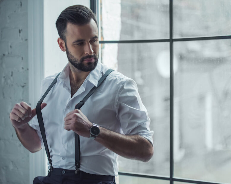 Handsome stylish man is holding his suspenders and looking away while sitting near the window