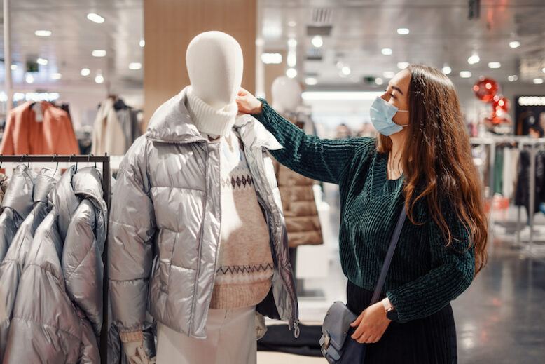 Young lady in face mask chooses warm clothes in department store, standing near mannequin dressed in puffy winter jacket