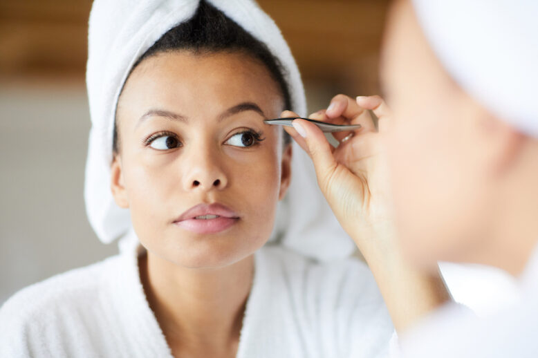Woman plucking eyebrows, looking in mirror during morning routine.
