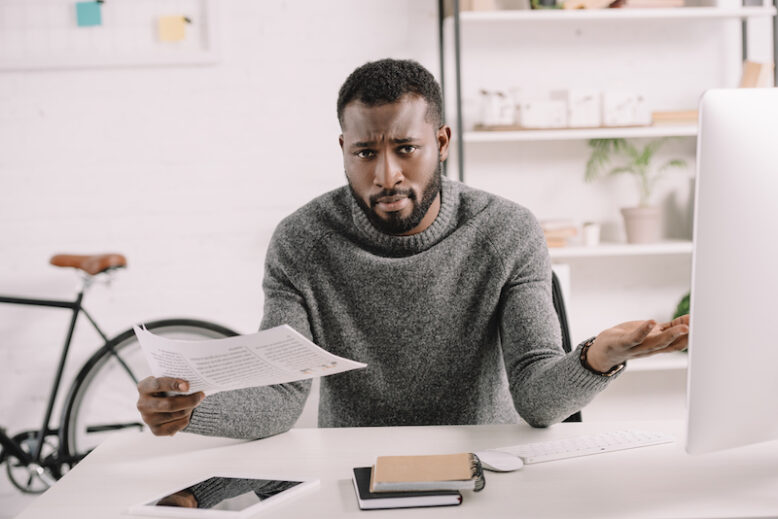 confused bearded african american businessman with shrug gesture holding document in office