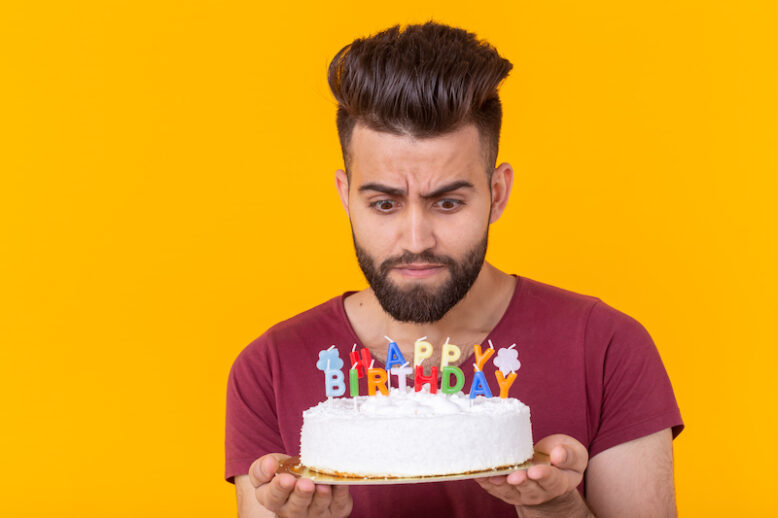 Embarrassed young male hipster with beard holding a birthday cake in his hands and looking thoughtfully at him posing on a yellow background.