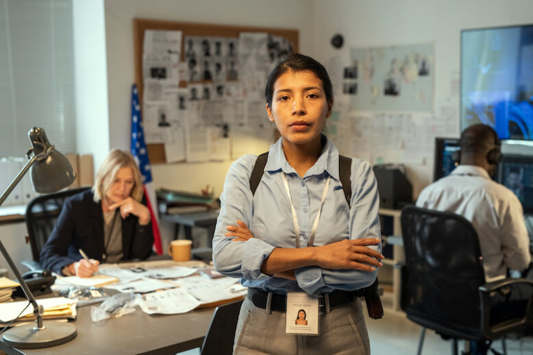 Young Hispanic female leader of intelligence service or police department in uniform crossing arms on chest while standing against coworkers