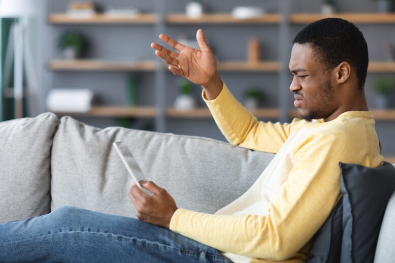 Angry african american young man holding digital tablet while reclining on couch in living room, gesturing, losing on trading markets, gambling online concept, side view, copy space