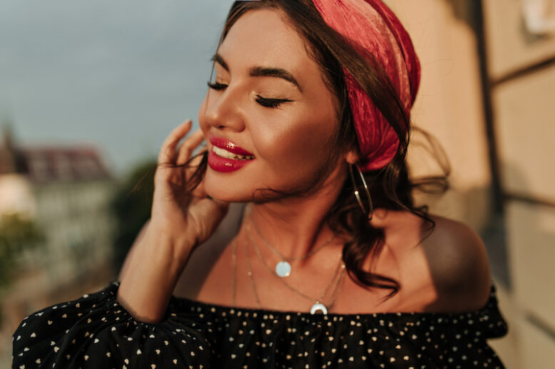 Cheerful tanned girl with big bright lips, accessories and brunette hair in fashionable dark polka dot blouse smiling and posing with closed eyes..