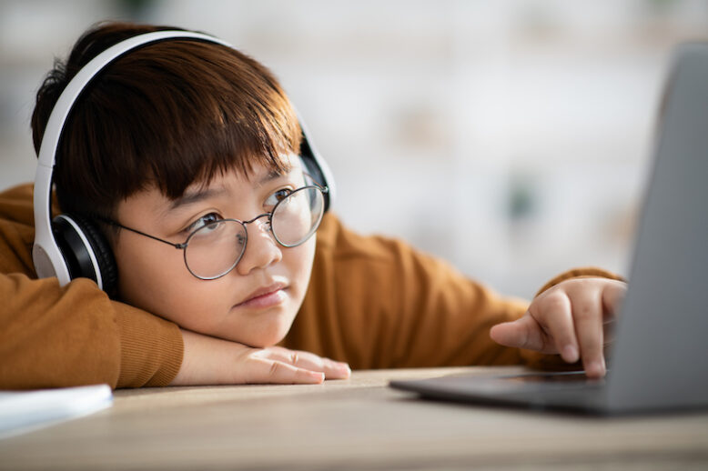 Closeup of bored plump chinese boy teenager wearing glasses studying online, sitting at desk at home, using laptop and wireless headset, attending class, copy space. Kids and lockdown concept