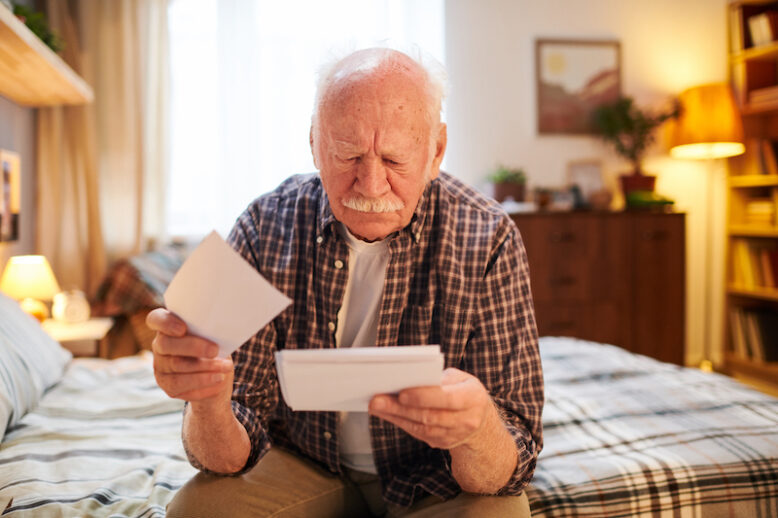 Old man sitting on the bed in bedroom and looking at photos in his hands, he remembering past events