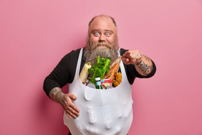 Surprised chunky bearded thick man indicates index finger at camera, has overweight as eats junk food, picks someone, stands against rosy background. Unhealthy nutrition and obesity concept.
