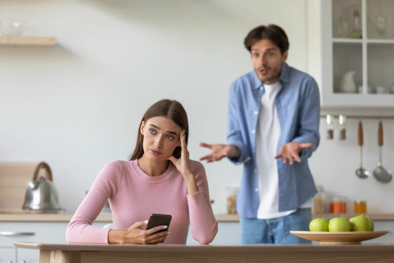 Young lady with phone ignoring frustrated scolding man in kitchen interior.