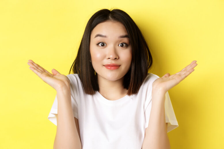 Close up of cute asian girl saying sorry, shrugging shoulders and smiling with oops face expression, standing over yellow background.