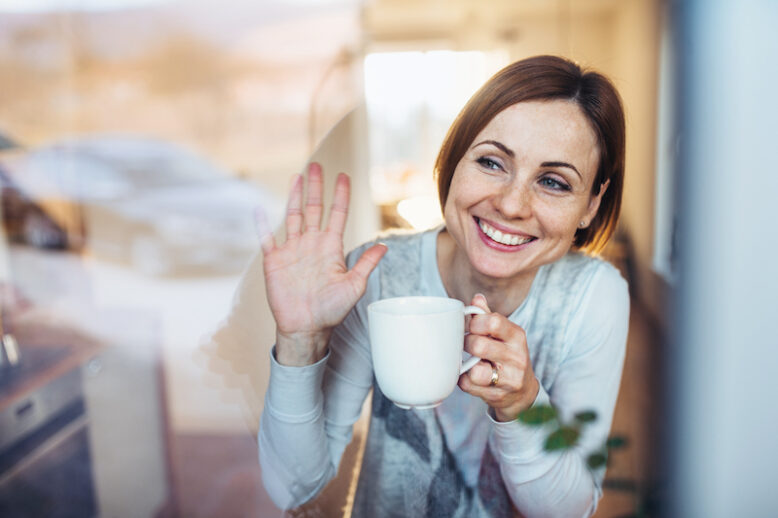A young woman with cup of coffee looking out of a window, waving goodbye to her husband in the morning.