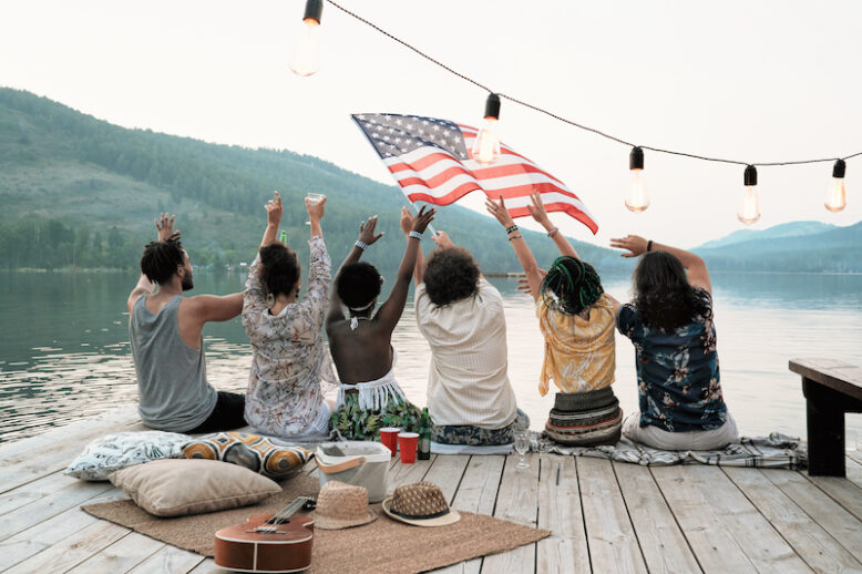 Rear view of young people sitting on a pier with American flag and celebrating the holiday on the nature
