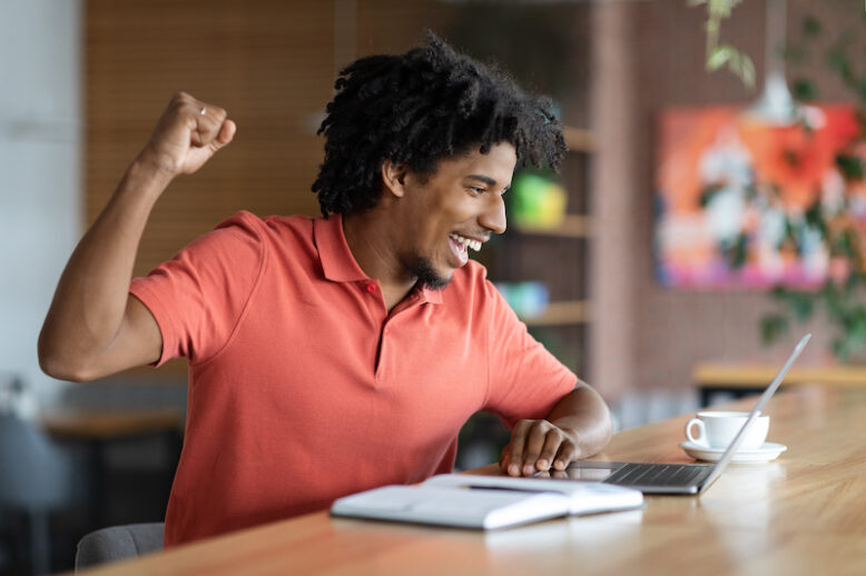 Good News. Emotional Black Man Looking At Laptop Screen And Celebrating Success While Sitting At Table In Cafe, Cheerful African American Guy Raising Fist And Exclaiming With Joy, Free Space
