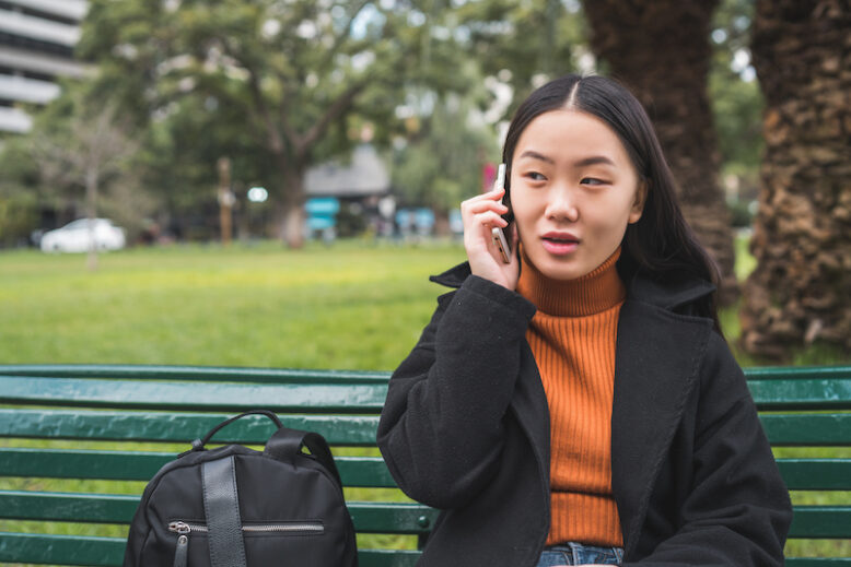 Portrait of young asian woman talking on the phone while sitting on a park bench.